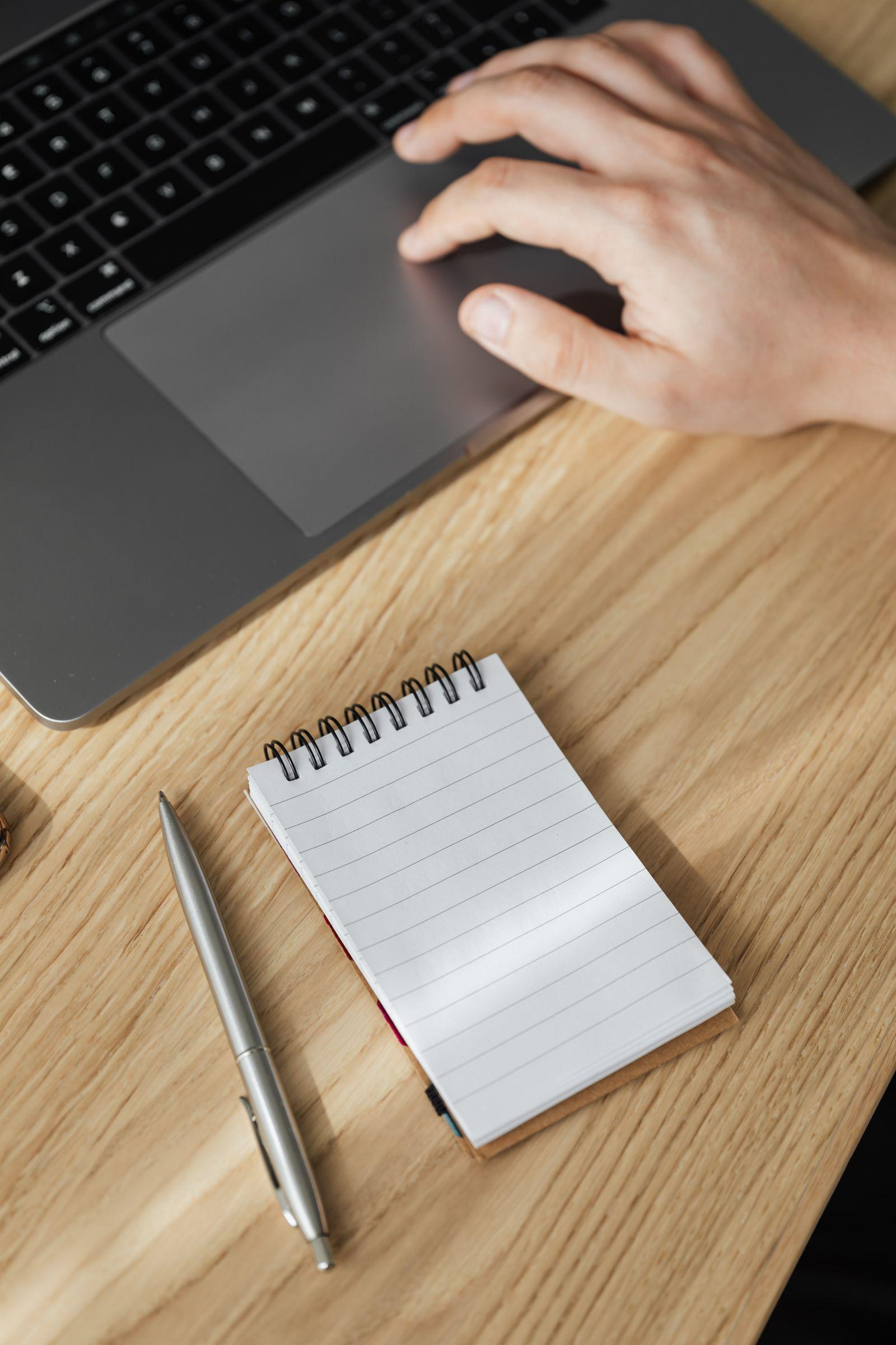 Dizüstü bilgisayarın touchpad'ine dokunan el, yanında spiralli defter ve kalem